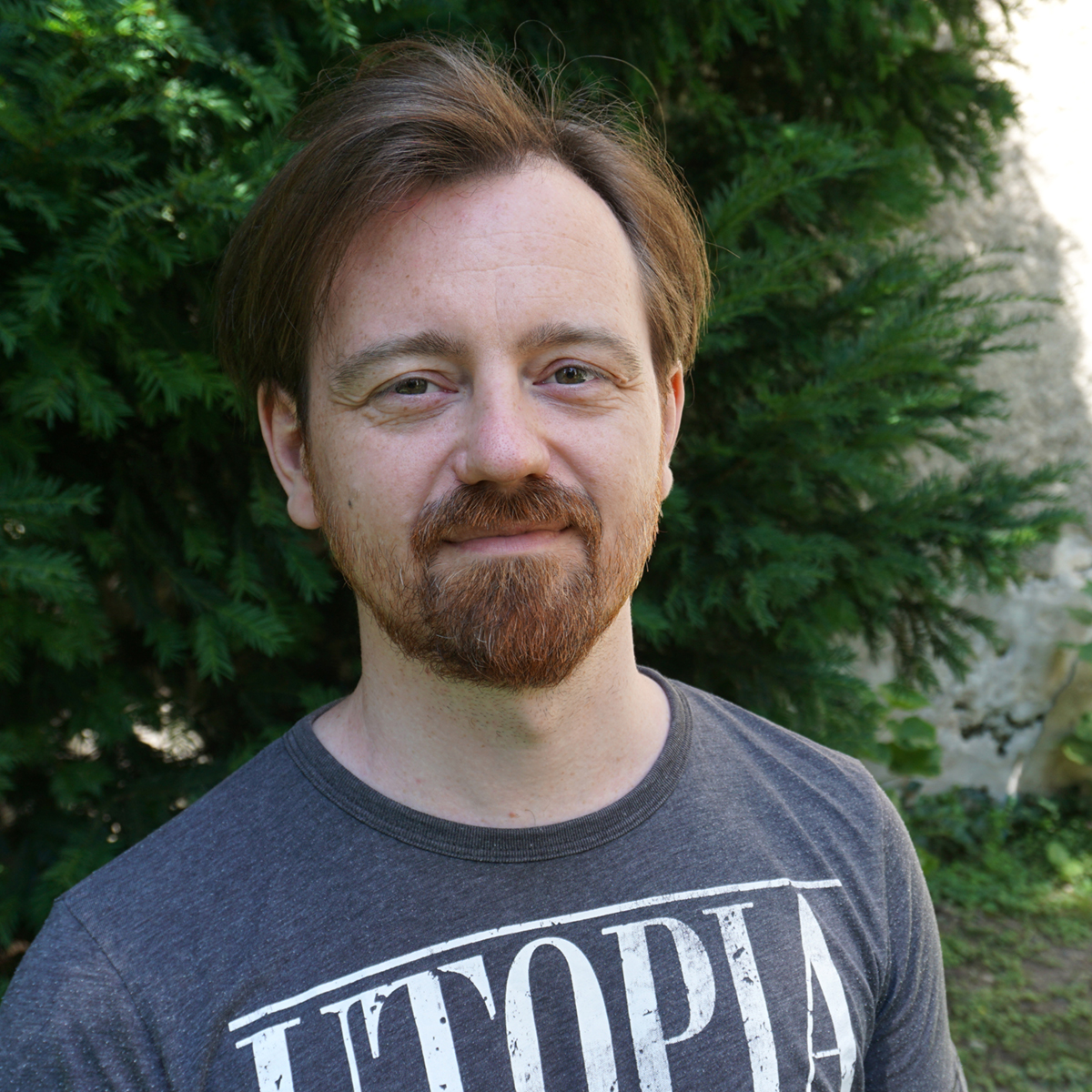 Ein Portrait von Alexander Schuc. Er lächelt in die Kamera, trägt ein graues Tshirt, hat hellbraune Haare und einen roten Bart. Im Hintergrund ist ein immergrüner Strauch zu sehen.