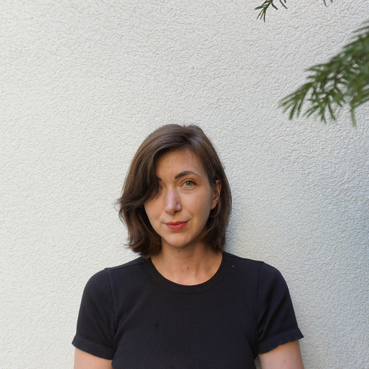 Ein Portrait von Hanna Helena Gerhardt. Sie schaut freundlich in die Kamera, hat braune kinnlange Haare und blasse Haut. Auf der rechten Seite ragt ein Ast eines immergrünen Strauchs in das Bild.